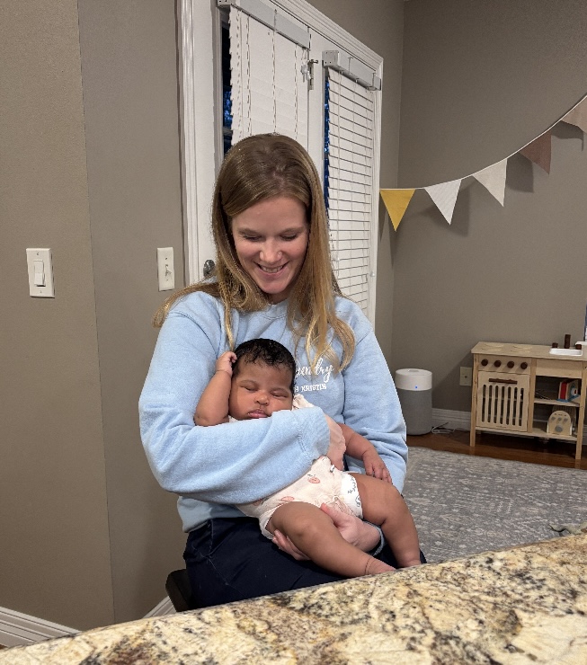 Kristin smiling as she holds a sleeping newborn in a nursery